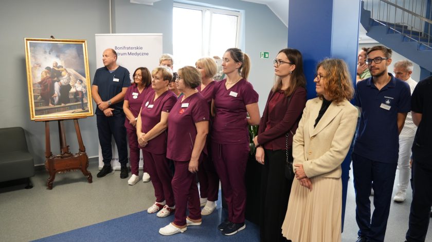 Grupa pielęgniarek w bordowych uniformach oraz lekarzy i rehabilitantów stoi na tle niebieskiej klatki schodowej, rozmawiając i uśmiechając się podczas spotkania w Bonifraterskim Centrum Medycznym