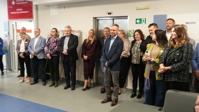 Grupa uczestników uroczystości w Bonifraterskim Centrum Medycznym stoi przed wejściem do Oddziału Rehabilitacji Szpitalnej. Wśród nich pracownicy, zaproszeni goście i przedstawiciele instytucji współpracujących