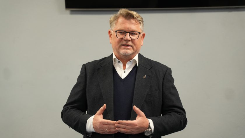 Mężczyzna w ciemnym garniturze przemawia podczas uroczystości w Bonifraterskim Centrum Medycznym, trzymając dłonie złożone przed sobą. W tle szara ściana i ekran