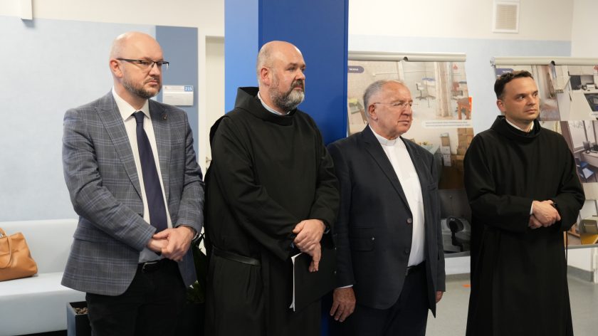 Trzej duchowni i mężczyzna w garniturze stoją obok plansz prezentujących zdjęcia sprzed i po remoncie szpitala. Wszyscy uważnie słuchają przemówienia