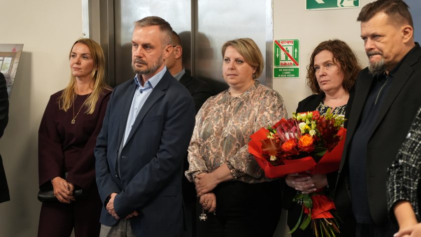 Uczestnicy wydarzenia stoją w skupieniu. Kobieta trzyma bukiet czerwonych i pomarańczowych kwiatów, obok niej mężczyźni w eleganckich strojach słuchają wystąpienia