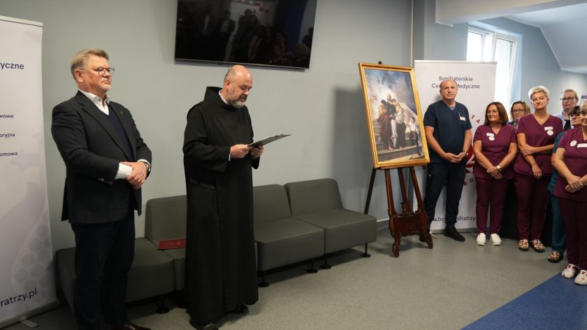 Grupa osób uczestniczy w uroczystości w Bonifraterskim Centrum Medycznym. Na pierwszym planie zakonnik w czarnej habicie odczytuje tekst z kartki. Obok stoi elegancko ubrany mężczyzna w garniturze. Po prawej stronie widać pielęgniarki w bordowych uniformach oraz obraz przedstawiający św. Jana Bożego niosącego chorego