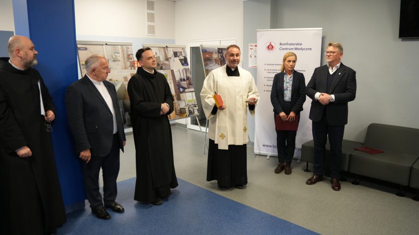Podczas uroczystości w Bonifraterskim Centrum Medycznym kilku zakonników i duchownych stoi przy ścianie. Jeden z nich, ubrany w białą albę, trzyma księgę i kropidło. Obok stoją przedstawiciele Centrum – kobieta w garniturze i mężczyzna w okularach. W tle widoczne są banery informacyjne o działalności placówki