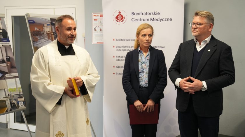 Zbliżenie na duchownego w albie, kobietę w granatowym garniturze oraz mężczyznę w okularach i ciemnej marynarce. W tle baner z logo Bonifraterskiego Centrum Medycznego i listą usług medycznych