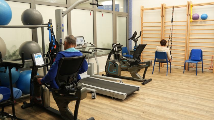 Sala rehabilitacyjna wyposażona w nowoczesny sprzęt: bieżnię, rowery treningowe, drabinki i piłki gimnastyczne. Pacjenci wykonują ćwiczenia pod okiem terapeutów