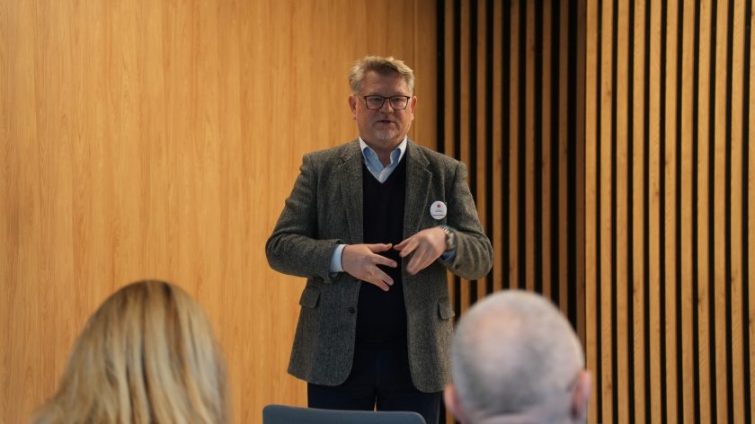 Mężczyzna w szarej marynarce mówi stojąc przed grupą słuchaczy. Za nim drewniana ściana, a na jego marynarce przypięta czerwona plakietka z logo Bonifratrów
