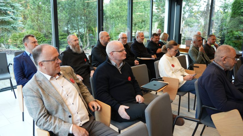 Grupa osób siedzi w jasnej sali konferencyjnej. Wśród uczestników widać kilku zakonników w habitach i osoby świeckie w eleganckich strojach
