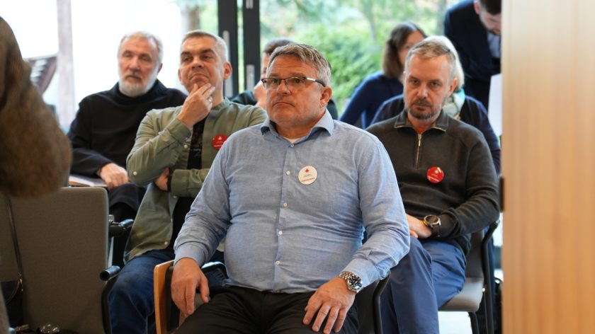 Uczestnicy spotkania siedzą w sali konferencyjnej, uważnie słuchając prelekcji. W pierwszym rzędzie mężczyzna w jasnoniebieskiej koszuli oraz dwaj inni uczestnicy z przypiętymi czerwonymi plakietkami Bonifraterskiego Centrum Medycznego. W tle widoczni zakonnicy i osoby świeckie w skupieniu