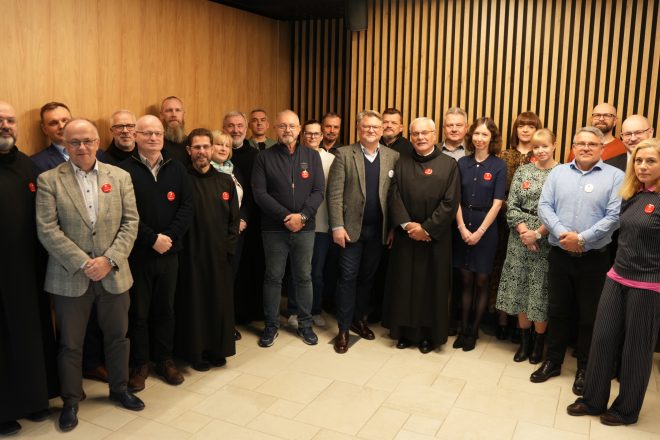 Wspólne zdjęcie grupowe uczestników spotkania — zakonnicy i świeccy stoją razem w sali z drewnianą ścianą. Wszyscy uśmiechnięci, z przypiętymi czerwonymi plakietkami