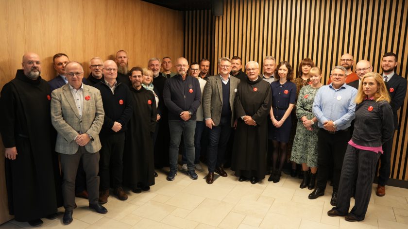 Wspólne zdjęcie grupowe uczestników spotkania — zakonnicy i świeccy stoją razem w sali z drewnianą ścianą. Wszyscy uśmiechnięci, z przypiętymi czerwonymi plakietkami