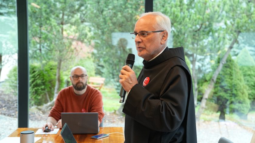 Zakonnik w habicie mówi do mikrofonu podczas wystąpienia w sali konferencyjnej. Za stołem z laptopem siedzi mężczyzna w czerwonym swetrze, a za nimi widać zieleń ogrodu przez duże okna