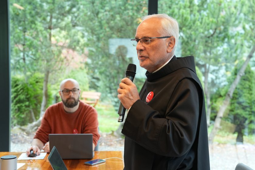 Zakonnik w habicie mówi do mikrofonu podczas wystąpienia w sali konferencyjnej. Za stołem z laptopem siedzi mężczyzna w czerwonym swetrze, a za nimi widać zieleń ogrodu przez duże okna