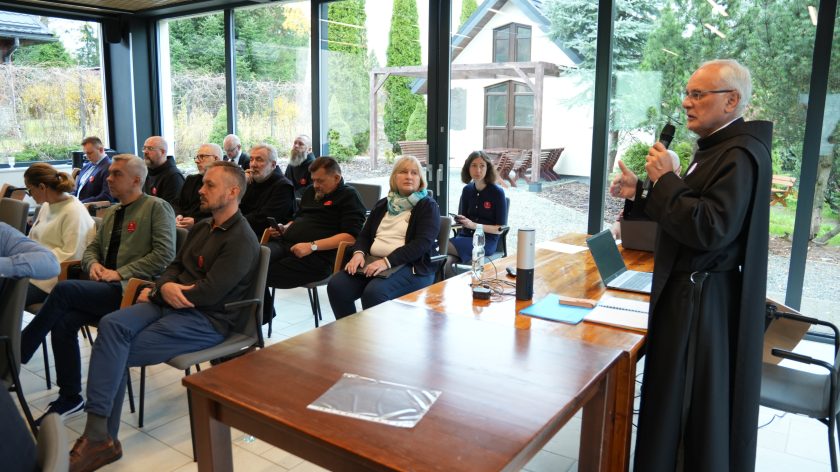 Spotkanie w sali konferencyjnej z dużymi oknami wychodzącymi na ogród. Przy stole stoi zakonnik w czarnym habicie i mówi do mikrofonu, prowadząc prezentację. Przed nim siedzi kilkanaście osób – zakonnicy i świeccy – uważnie słuchający prelekcji