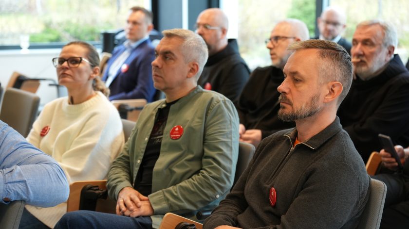 Uczestnicy spotkania siedzą w sali konferencyjnej i z uwagą słuchają wystąpienia. W pierwszym rzędzie kobieta w białym swetrze oraz mężczyźni w ciemnych ubraniach z przypiętymi czerwonymi plakietkami Bonifraterskiego Centrum Medycznego