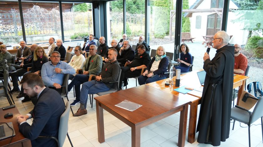 Spotkanie w sali konferencyjnej z dużymi oknami wychodzącymi na ogród. Przy stole stoi zakonnik w czarnym habicie i mówi do mikrofonu, prowadząc prezentację. Przed nim siedzi kilkanaście osób – zakonnicy i świeccy – uważnie słuchający prelekcji