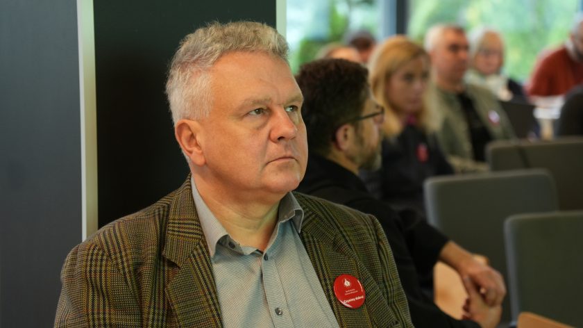 Mężczyzna w koszuli i marynarce z uwagą spogląda na wyznaczony punkt. W tle widać inne osoby uczestniczące w wydarzeniu, konferencji . Mężczyzna na pierwszym planie ma plakietkę Bonifraterskiego Centrum Medycznego z hasłem: Czynimy dobro!