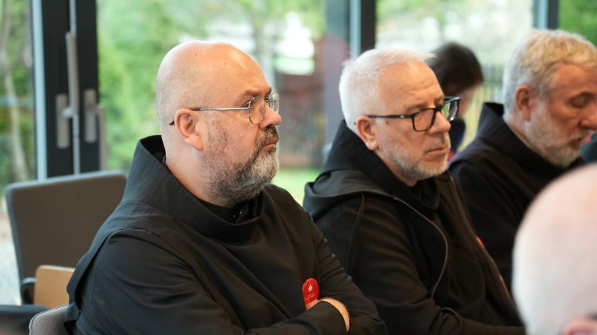 Trzech zakonników w czarnych habitach siedzi w sali konferencyjnej. Mężczyźni słuchają z powagą, w tle widoczne duże okna z widokiem na ogród