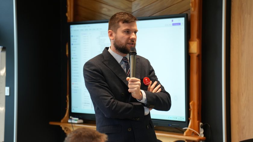 Mężczyzna w granatowym garniturze mówi do mikrofonu podczas spotkania. Za nim ekran z prezentacją, a na klapie marynarki przypięta czerwona plakietka z napisem „Czynimy dobro!”