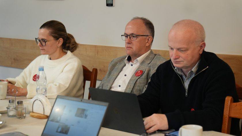 Dwóch mężczyzn i kobieta w eleganckich strojach siedzą przy stole i pracuje przy laptopach. Na stole filiżanka, butelka wody i dokumenty. Uczestnicy skupieni na omawianiu materiałów podczas spotkania roboczego