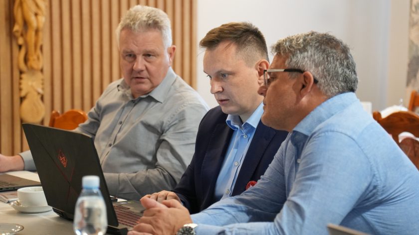 Trzech mężczyzn w eleganckich strojach siedzi przy stole i pracuje przy laptopach. Na stole filiżanka, butelka wody i dokumenty. Uczestnicy skupieni na omawianiu materiałów podczas spotkania roboczego