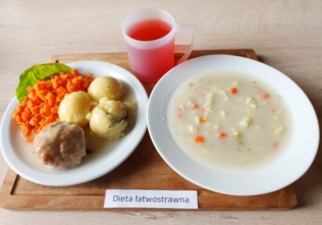 Na drewnianej tacy znajduje się zestaw obiadowy dla osób wymagających diety łatwostrawnej. Na talerzu ułożono gotowaną marchewkę w kostkę, kilka ziemniaków z koperkiem oraz pulpet w jasnym sosie. Obok stoi kubek z czerwonym kompotem. Po prawej stronie widoczny jest talerz z lekką zupą jarzynową z makaronem i kawałkami marchewki