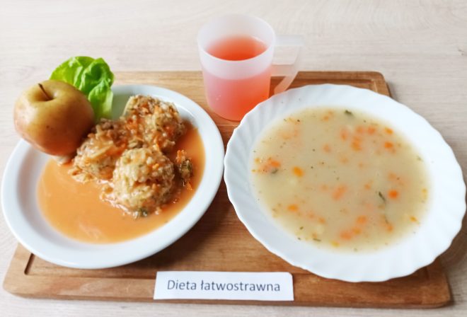 Obiad: pulpety ryżowo-mięsne w sosie pomidorowym, podane z jabłkiem i liściem sałaty. Obok zupa jarzynowa w talerzu oraz kubek z kompotem. Na stoliku kartka z napisem Dieta łatwostrawna