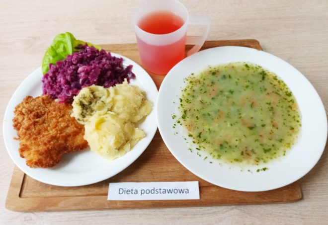 Obiad: kotlet schabowy w panierce z ziemniakami posypanymi koperkiem i surówką z czerwonej kapusty. Obok talerz z zupą jarzynową i kubek z czerwonym kompotem. Na stoliku kartka z napisem Dieta podstawowa