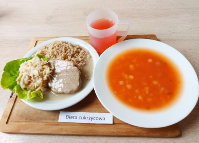 Na drewnianej tacy zestaw obiadowy opisany jako „Dieta cukrzycowa”. Na talerzu znajduje się porcja kaszy, kawałek duszonego mięsa w jasnym sosie oraz surówka z białej kapusty i marchwi podana na liściu sałaty. Obok stoi talerz z zupą pomidorową z ziemniakami oraz kubek z czerwonym napojem owocowym