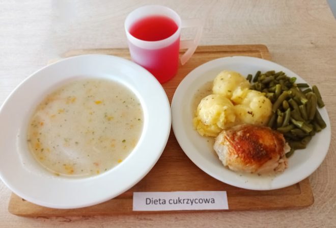 Na tacy z podpisem „Dieta cukrzycowa” znajduje się zestaw obiadowy składający się z trzech elementów: po lewej stronie talerz z jasną zupą jarzynową na bazie wywaru warzywnego z widocznymi kawałkami marchwi i koperku; po prawej stronie drugi talerz z porcją gotowanych ziemniaków, duszoną zieloną fasolką szparagową oraz kawałkiem pieczonego kurczaka w sosie; na górze plastikowy kubek z czerwonym napojem owocowym bez dodatku cukru. Całość ustawiona jest na drewnianej deseczce