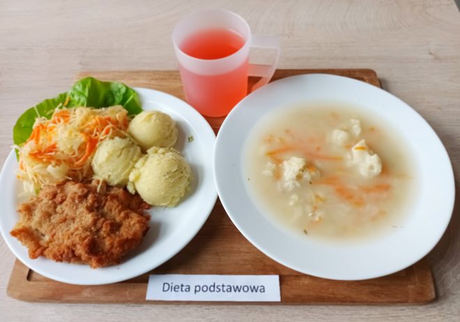 Taca z obiadem oznaczona etykietą „Dieta podstawowa”. Na talerzu znajduje się kotlet panierowany, dwie porcje puree ziemniaczanego oraz surówka z kiszonej kapusty i marchewki ułożona na liściu sałaty. Obok stoi miska z zupą kalafiorową z dodatkiem marchewki oraz kubek z czerwonym napojem owocowym
