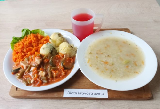 Zestaw obiadowy w diecie łatwostrawnej. Na talerzu ziemniaki z koperkiem, gulasz warzywny z mięsem drobiowym oraz surówka z gotowanej marchewki na liściu sałaty. Obok talerza miska z zupą jarzynową oraz kubek z czerwonym napojem. Na tabliczce widoczny napis „Dieta łatwostrawna”