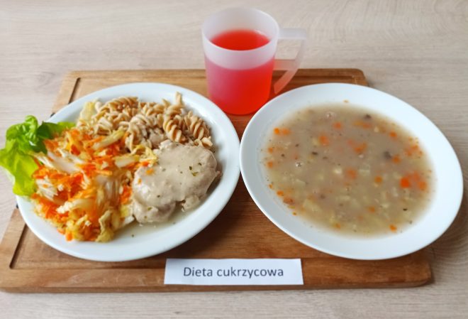 Taca z posiłkiem oznaczonym jako „Dieta cukrzycowa”. Na talerzu znajduje się porcja pełnoziarnistego makaronu, gotowane mięso w jasnym sosie, surówka z kapusty i marchewki oraz liść sałaty. Obok talerza stoi miska z zupą jarzynową z kawałkami marchwi, ziemniaków i kaszy. Do posiłku podano kubek z czerwoną herbatą owocową
