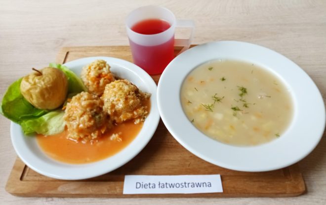 Zestaw obiadowy w diecie łatwostrawnej. Na tacy stoją dwa białe talerze: na jednym widoczna jest jasna zupa jarzynowa z koperkiem, na drugim trzy pulpety ryżowo-mięsne w lekkim sosie pomidorowym, obok pieczone jabłko na liściu sałaty. Do posiłku podany czerwony kompot w przezroczystym kubku z uchem
