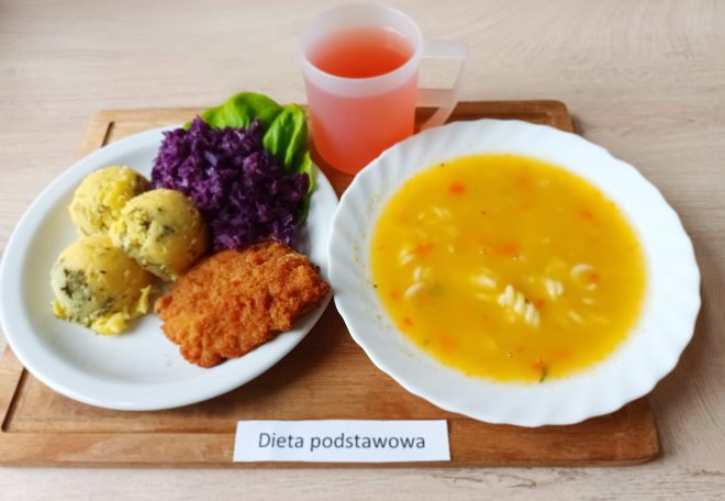 Zestaw obiadowy z zupą jarzynową z makaronem oraz drugim daniem: panierowany kotlet, ziemniaki z koperkiem, surówka z czerwonej kapusty i liść sałaty. Do posiłku podano różowy napój w plastikowym kubku. Na kartce napis: „Dieta podstawowa”