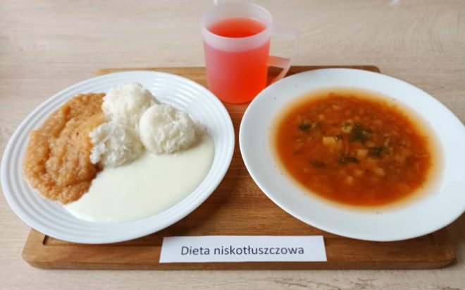 Na drewnianej tacy ustawiono posiłek opisany etykietą „Dieta niskotłuszczowa”. Na jednym talerzu znajdują się trzy porcje ryżu, sos jogurtowy lub mleczny oraz przecier z gotowanych jabłek. Obok podano talerz z zupą jarzynową o czerwonej barwie, z kawałkami warzyw i koperkiem. Do zestawu dołączony jest kubek z czerwonym napojem owocowym. Całość wygląda lekko i dietetycznie