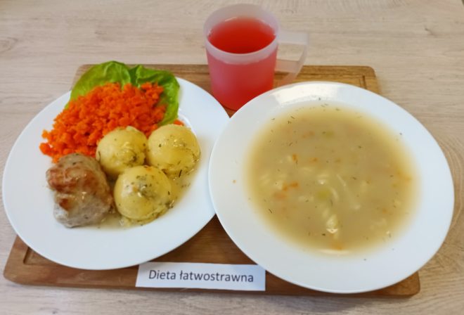 Na tacy leży posiłek oznaczony jako „Dieta łatwostrawna”. Na talerzu znajdują się trzy ziemniaki polane sosem, kawałek pieczonego mięsa oraz porcja gotowanej, rozdrobnionej marchewki z listkiem sałaty. Obok stoi miska z jasną zupą jarzynową oraz kubek z czerwonym kompotem lub napojem owocowym
