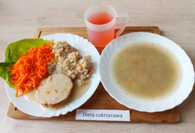 Zestaw obiadowy dla diety cukrzycowej: talerz z gotowaną kaszą, sosem i plasterkiem mięsa, surówką z marchwi oraz listkiem sałaty. Obok zupa jarzynowa i różowy napój w plastikowym kubku. Posiłek ustawiony na drewnianej tacy