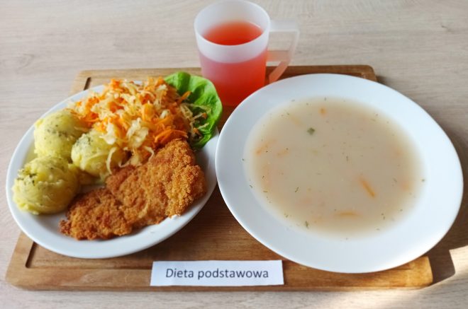 Tacka z obiadem w diecie podstawowej. Na talerzu panierowany filet z ryby z ziemniakami posypanymi koperkiem i surówką z kapusty i marchwi. Obok talerz z jasną zupą warzywną oraz kubek z czerwonym napojem owocowym