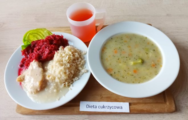 Obiad w diecie cukrzycowej podany na tacy. Na talerzu danie główne: pierś z kurczaka w sosie z kaszą i buraczkami. Obok talerz z zupą brokułową i kubek czerwonego napoju