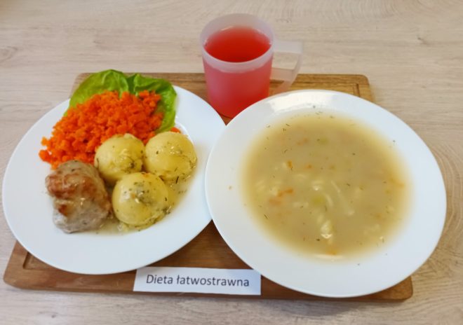 Zestaw obiadowy w diecie łatwostrawnej. Na tacy talerz z zupą jarzynową, obok danie główne: ziemniaki z koperkiem, duszone mięso i marchewka z sałatą. Do posiłku czerwony napój w plastikowym kubku