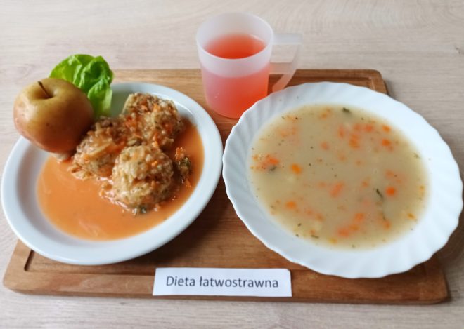 Na tacy z napisem „Dieta łatwostrawna” znajduje się miska z zupą jarzynową o jasnym kolorze oraz talerz z pulpetami w sosie pomidorowym, liściem sałaty i jabłkiem. Obok stoi kubek z czerwonym napojem owocowym