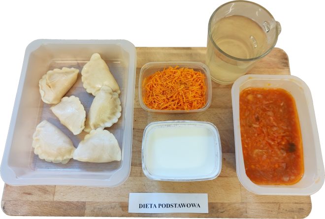 Obiad: porcja pierogów w plastikowym pojemniku, surówka z marchwi, zupa jarzynowa w czerwonym sosie oraz kubek z jasnym napojem. Dodatkowo mały kubeczek jogurtu naturalnego. Na stoliku kartka z napisem Dieta podstawowa