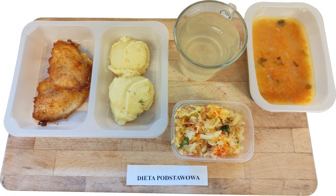 Na tacy ustawiono pojemnik z panierowanym kotletem drobiowym i ziemniaczanym puree. Obok porcja surówki z kiszonej kapusty, talerz z zupą jarzynową oraz kubek z jasnym napojem. Całość opisana etykietą „Dieta podstawowa”