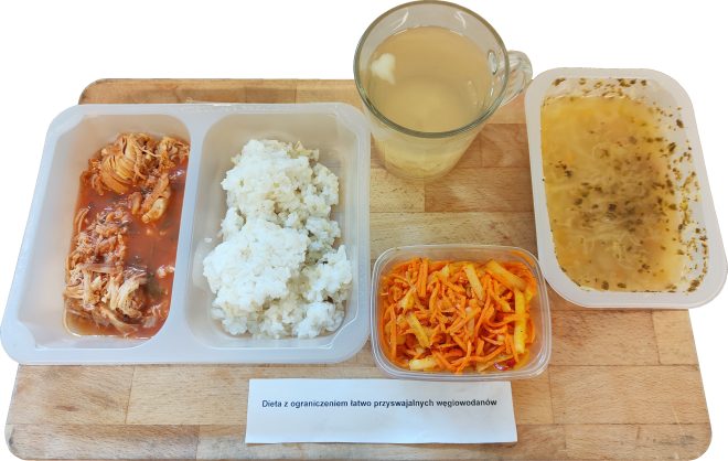 Na drewnianej tacy znajduje się zestaw obiadowy w pojemnikach dietetycznych opisany jako „Dieta z ograniczeniem łatwo przyswajalnych węglowodanów”. W skład posiłku wchodzi porcja gotowanego ryżu, duszone mięso drobiowe w sosie pomidorowym, zupa w przezroczystym pojemniku ziołowym, surówka z marchewki i selera oraz kubek z napojem przypominającym kompot lub herbatę owocową. Całość podana jest w estetycznej formie szpitalnej diety lekkostrawnej