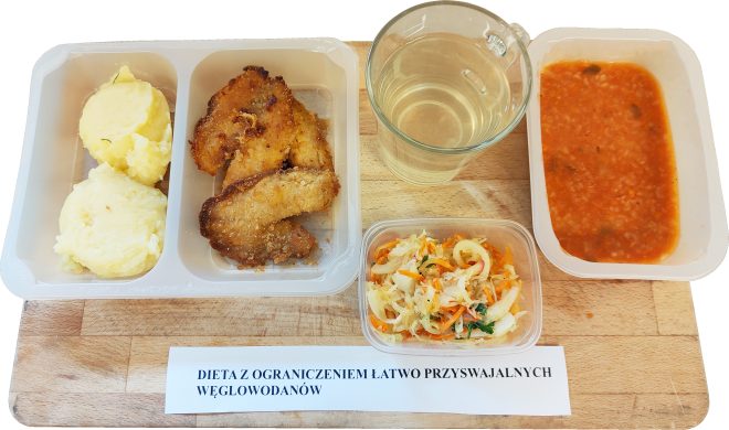 Zestaw obiadowy w pojemnikach dietetycznych opisany jako „Dieta z ograniczeniem łatwo przyswajalnych węglowodanów”. W skład posiłku wchodzą puree ziemniaczane, panierowane kawałki mięsa, surówka z kapusty i marchwi, zupa pomidorowa z ryżem oraz szklanka jasnego napoju. Całość ustawiona na drewnianej desce