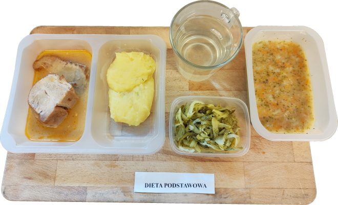 Na tacy z napisem „Dieta podstawowa” znajduje się zestaw obiadowy w pojemnikach. W pierwszej przegródce podano dwa kawałki gotowanego mięsa w sosie, w drugiej porcję puree ziemniaczanego. Obok znajduje się surówka z duszonej kapusty z koperkiem, szklanka wody oraz zupa jarzynowa z warzywami takimi jak marchew, ziemniaki i brokuły. Całość ułożona jest na drewnianej desce