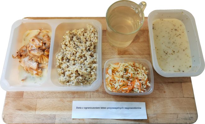 Zestaw obiadowy podany w pojemniku cateringowym. W jednym przedziale znajduje się mięso w sosie z podsmażoną cebulą, w drugim kasza jęczmienna. Obok stoi przezroczysty kubek z jasnym napojem, sałatka z białej kapusty i marchewki w osobnym pojemniku oraz miska z zupą kremem. Całość ustawiona na drewnianej desce, z karteczką z napisem „Dieta z ograniczeniem łatwo przyswajalnych węglowodanów”
