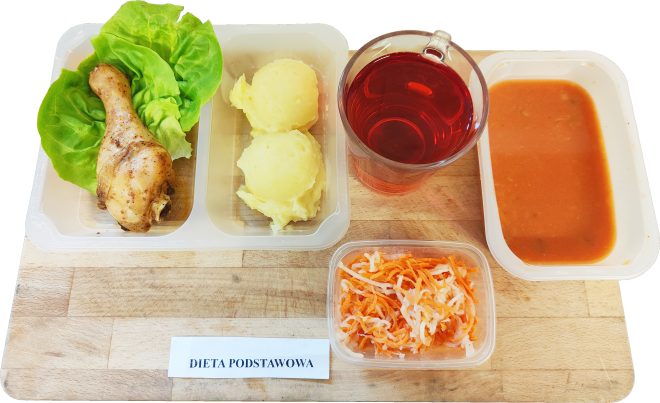 Na tacy znajduje się zestaw obiadowy złożony z gotowanego podudzia kurczaka ułożonego na liściach sałaty, dwóch porcji puree ziemniaczanego, czerwonego napoju w szklance, surówki z marchewki i białej kapusty oraz zupy pomidorowej w pojemniku. Etykieta informuje: „Dieta podstawowa”