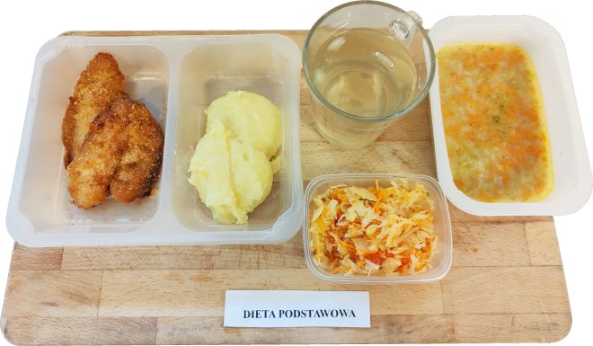 Na tacy znajduje się zestaw obiadowy w diecie podstawowej: panierowany filet z kurczaka, puree ziemniaczane, surówka z białej kapusty i marchewki, zupa jarzynowa oraz szklanka z jasnym napojem. Etykieta informuje: „Dieta podstawowa”