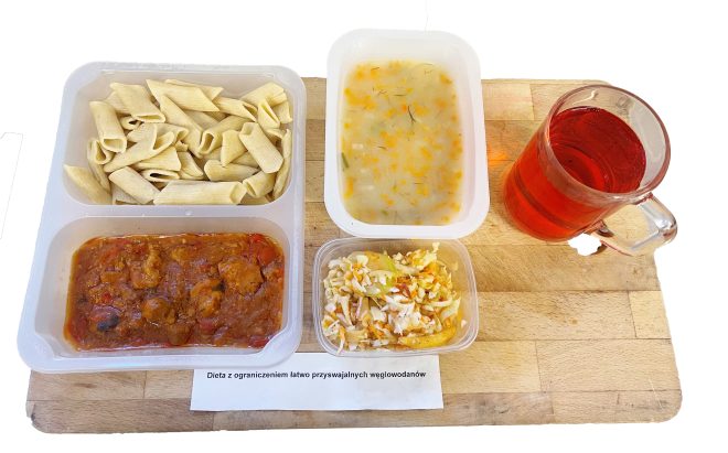 Na drewnianej desce ułożony jest zestaw obiadowy w plastikowych pojemnikach, opisany etykietą „Dieta z ograniczeniem łatwo przyswajalnych węglowodanów”. W jednym z pojemników znajdują się rurki makaronu pełnoziarnistego, w drugim danie mięsno-warzywne w sosie pomidorowym. Obok znajduje się porcja zupy jarzynowej z marchewką i koperkiem oraz niewielki pojemnik z surówką z kapusty, marchewki i ogórka. Do zestawu dołączony jest kubek z czerwonym napojem owocowym